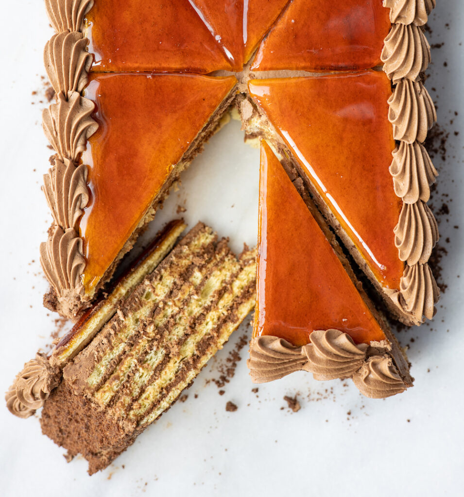 an overhead view of a dobos torta with a slice pulled slightly out and turned on its side to reveal the cake's layers
